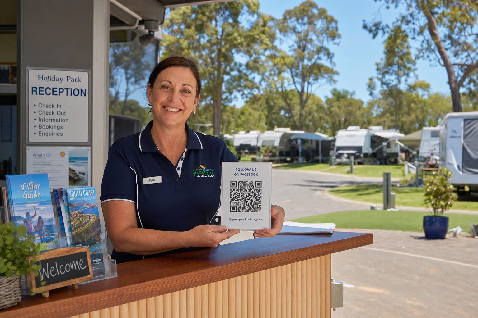 Friendly caravan park host at reception holding a QR referral code, with caravans on grassy sites behind
